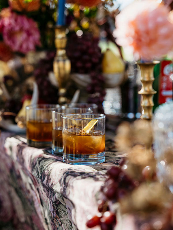 Three glasses of amber-colored cocktails with a lemon twist on a decorative table with flowers and fruits.