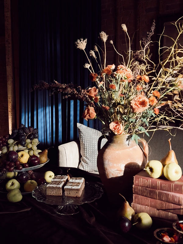 Decorative table setting with flowers, fruits, and books against a dark curtain.
