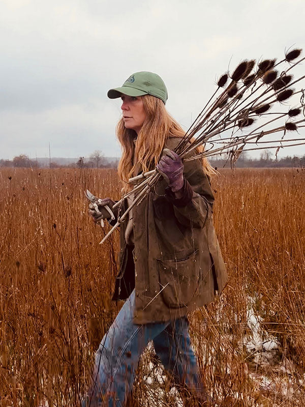 Christine holding dried plants in a field wearing a green cap and jacket.