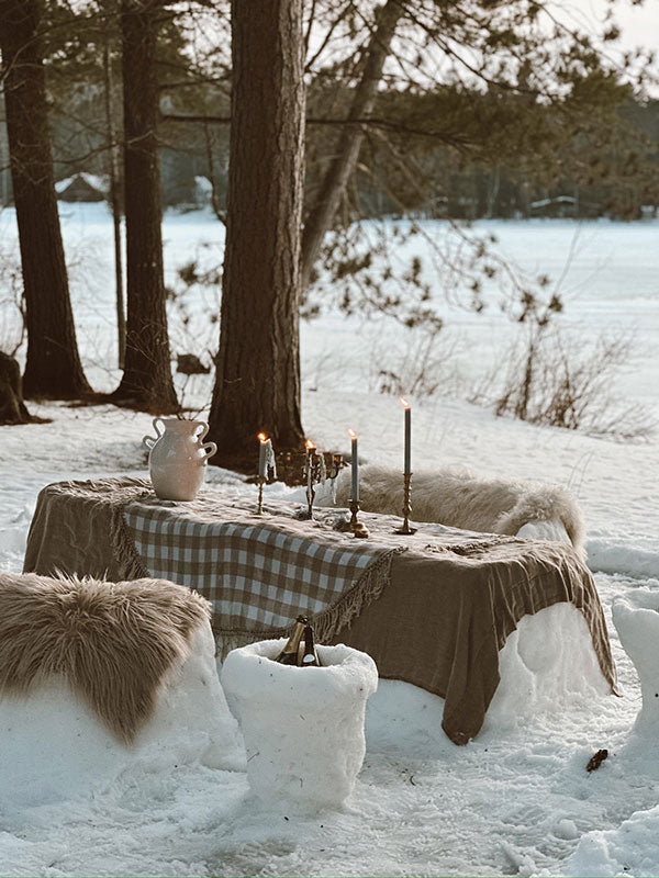 Dining table set in a snowy outdoor setting with candles and a checkered tablecloth.