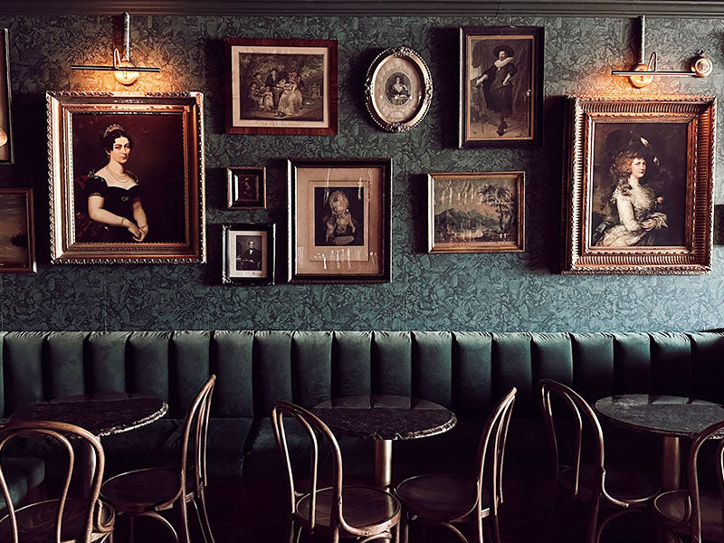 Vintage-style interior with framed portraits on a textured wall above a row of tables and chairs.